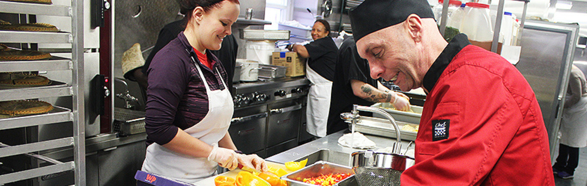 Chef Ric Watson testing the temperature of a meal with a thermometer while a trainee works behind him